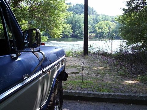 1978 Ford Bronco - Close view down the driver's side of a blue and silver two-tone Ford truck parked facing a wooded river, with the door mirror, hood edge an…