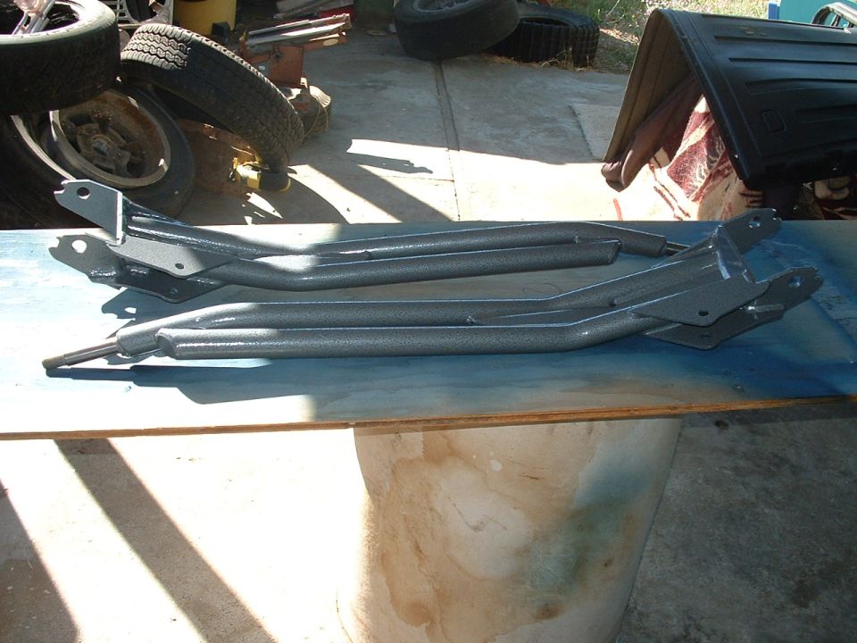 Two gray metal suspension parts laid out on a blue surface, with tires and other items in the background