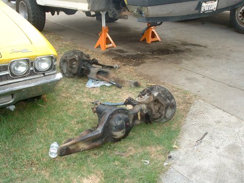 1990 Ford Bronco - Vehicle axles are laid out on the grass, with a yellow car and a white vehicle elevated on jack stands in the background.