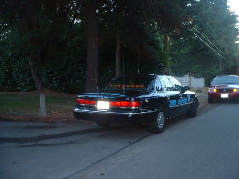 1999 Ford Crown Victoria - A black sedan, likely a 1998 model, is parked on the side of the road with antennas and a light bar. A second vehicle is visible ahead.