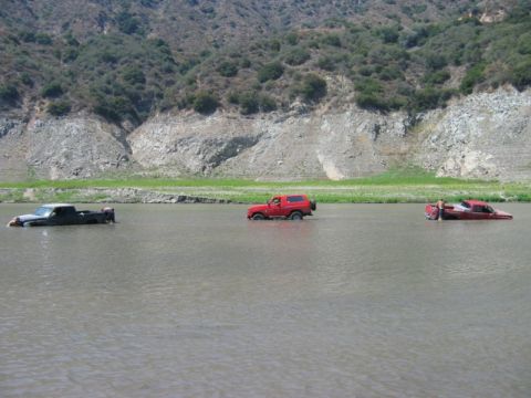 1985 Ford Bronco - A red 1985 Ford Bronco navigates through a flooded area, accompanied by two trucks partially submerged in the water.