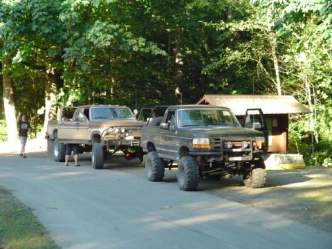 1993 Ford Bronco - Two heavily modified trucks are parked on a road near a small building in a wooded area.