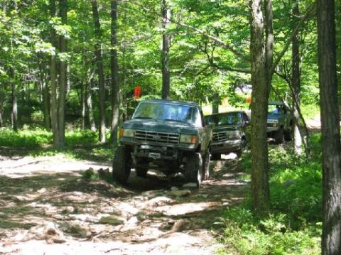 1995 Ford Bronco - Multiple off-road vehicles navigate a wooded trail with rocky terrain.