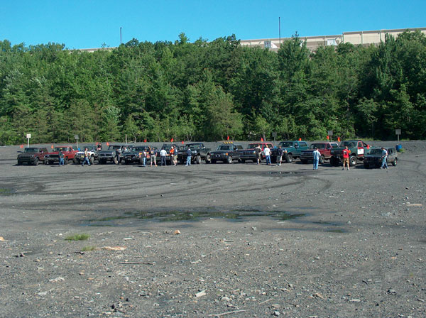 Row of trucks and SUVs with whip flags parked along a gravel lot edged by pine trees
