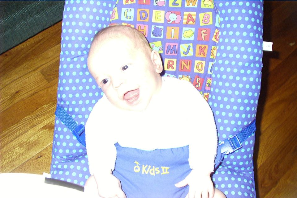Baby in a blue polka dot bouncer with alphabet blocks pattern on the back. Text 'Kids II' visible on the baby's clothing