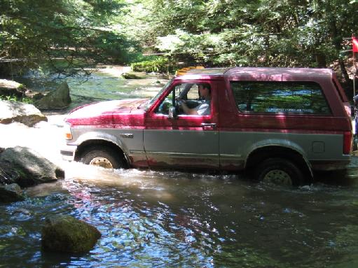 Maroon and silver Ford Bronco fording a rocky shallow creek in the woods