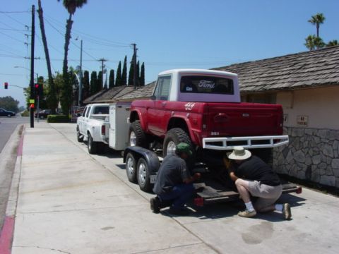 1992 Ford Bronco - A red Ford Bronco is being loaded onto a trailer towed by a white pickup truck, with two individuals attending to the rear of the Bronco.