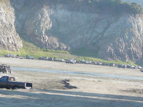 1992 Ford Bronco - Multiple vehicles are parked on a dirt lot with a large rocky cliff face behind them.