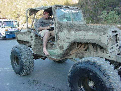 1992 Ford Bronco - A heavily mud-caked off-road vehicle sits on pavement next to a tow truck.