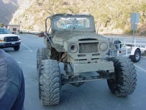 1992 Ford Bronco - A heavily mud-caked off-road vehicle sits parked on asphalt, with a white pickup truck visible in the background.