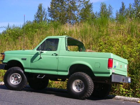 1990 Ford Bronco - Rear three-quarter driver-side view of a lifted mint-green Ford Bronco with the hardtop removed, showing the open rear tub and tan interior…