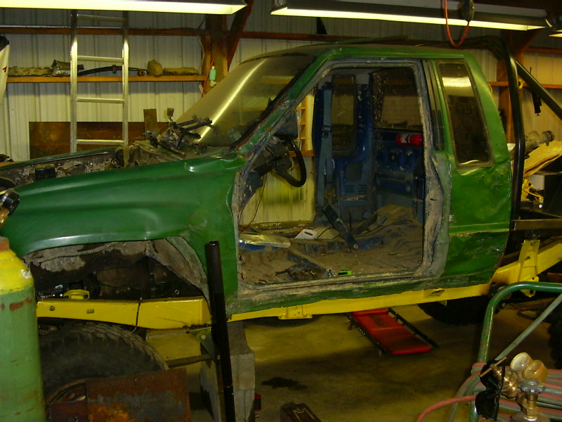 Green pickup truck chassis with yellow frame, missing doors and fenders, in a workshop
