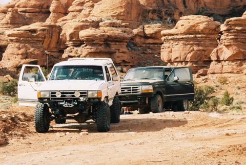 1996 Ford Bronco - Two Ford Broncos are parked on a dirt road in a desert landscape with red rock formations.