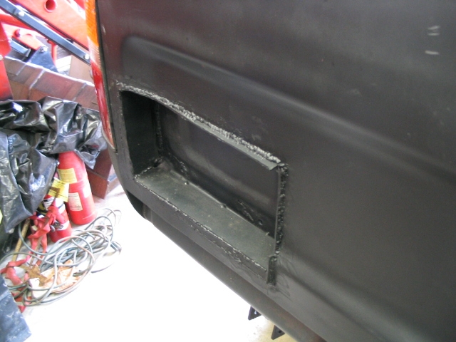 Black panel with a rectangular cutout, showing welded metal inside. Wires and fire extinguishers visible to the left