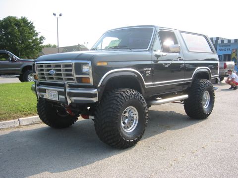 1996 Ford Bronco - This lifted 1980s Ford Bronco features prominent chrome trim and oversized off-road tires, parked on pavement with another vehicle visible…