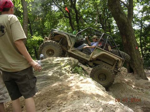 1992 Ford Bronco - A modified off-road vehicle navigates a challenging rocky terrain on a wooded trail.