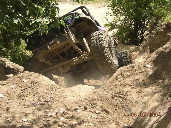 Off-road vehicle with large tires climbing a steep, dusty dirt hill with rocks and trees