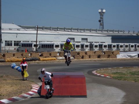 1990 Ford Bronco - Two riders compete on electric scooters, one airborne over a red ramp, with buses and a tower in the background.