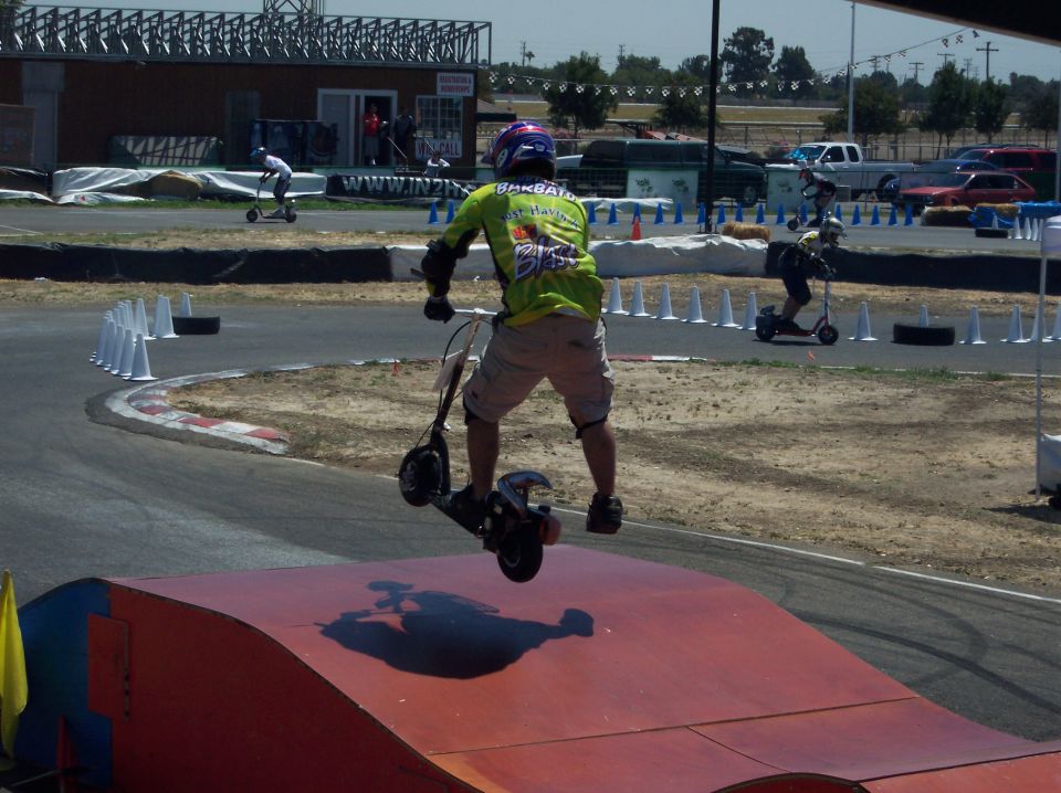 Person on a scooter mid-air over a ramp at a race track, with other riders and vehicles in the background