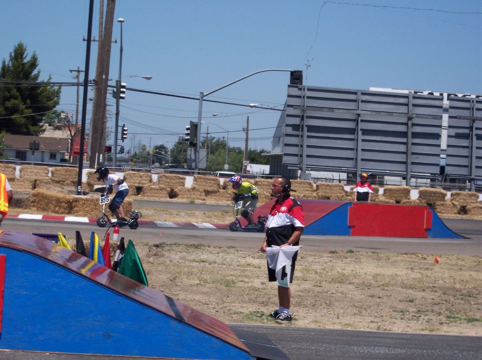 Scooter race with riders on track, officials nearby, hay bales, and ramps