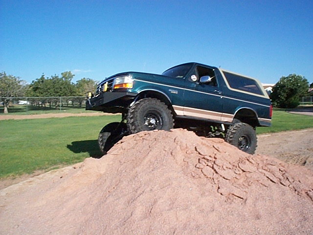 Lifted green-and-tan 1990s Ford Bronco parked on top of a reddish dirt mound under blue sky