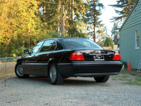 1999 BMW 7 Series - The rear of a black 1999 BMW 7 Series sedan is visible, parked on a gravel driveway next to a house.
