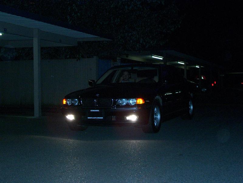 Dark BMW sedan at night with lights on, front 3/4 view in a carport lot