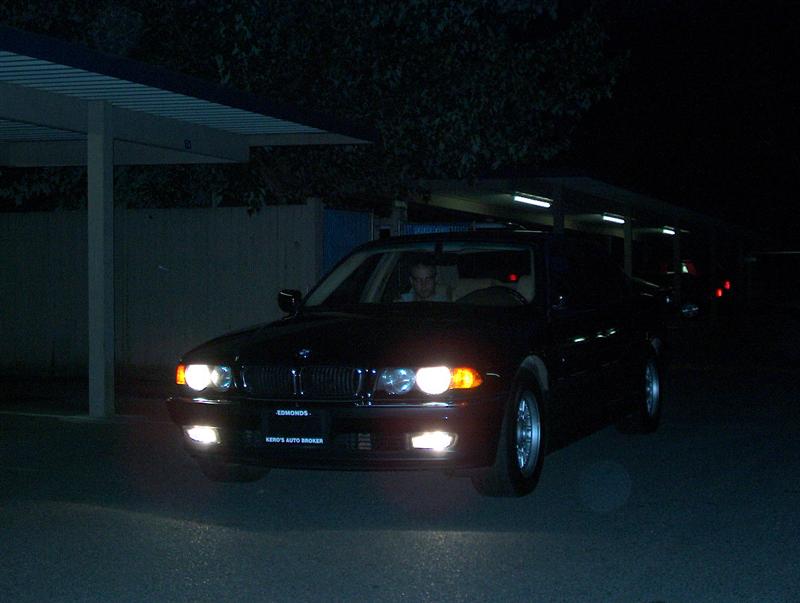 Dark BMW sedan at night with headlights on, driver inside, near a carport.