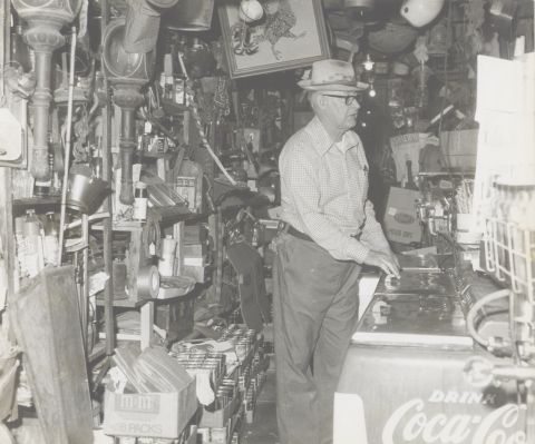 1995 Ford Bronco - A man stands behind a counter in a cluttered store, surrounded by merchandise.