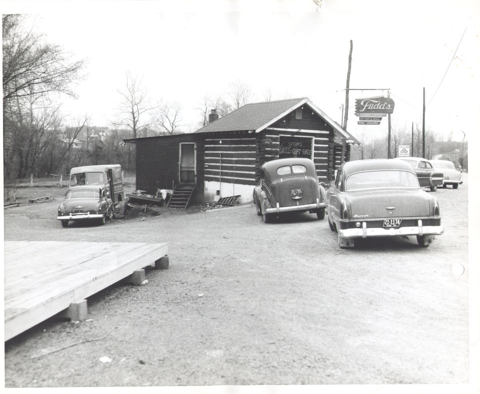 Log cabin building with 'Fudd's' sign, parked cars including a sedan and a van