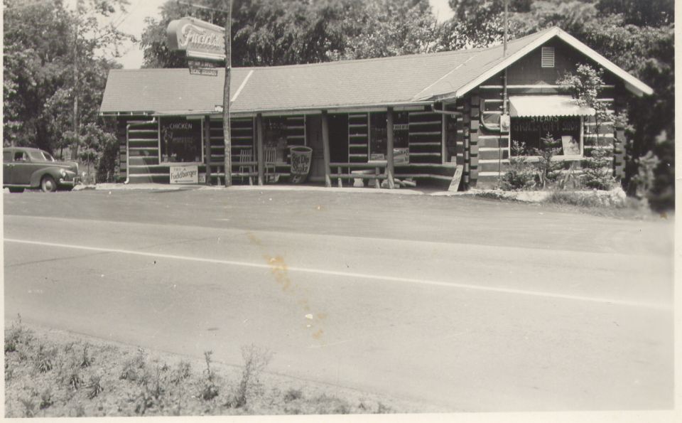 Log cabin building with a sign that says 'Fuddtown'. A car is parked to the left. Road in foreground