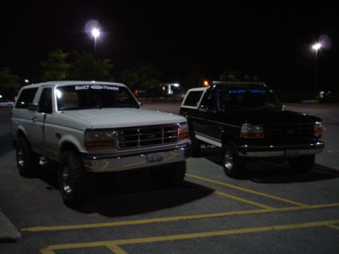1993 Ford Bronco - Two Ford Broncos, one white and one black, are parked side-by-side in a lot at night.