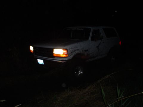 1993 Ford Bronco - A white SUV is seen at night with its headlights illuminated, covered in mud.