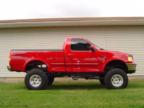 1978 Ford Bronco - Passenger-side profile of a red regular-cab short-bed Ford F-150 pickup on a suspension lift, parked on grass in front of a beige metal bui…
