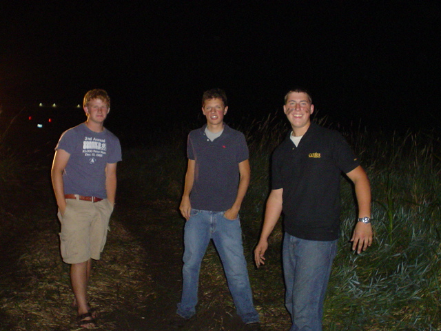 Three young men stand in a grassy area at night, with lights visible in the distance