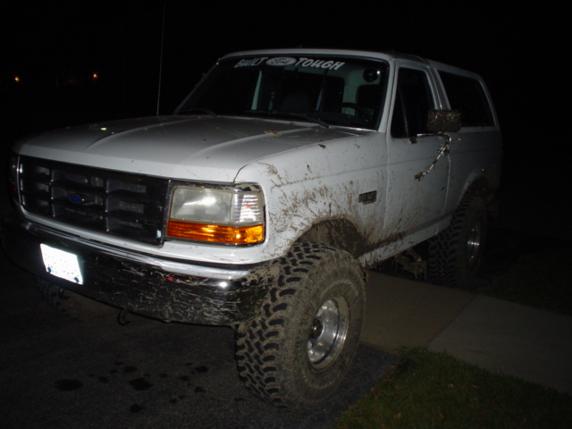 Mud-splattered white Ford Bronco with off-road tires. Banner on windshield reads 'BUILT FORD TOUGH'