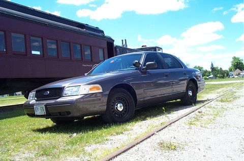 2001 Ford Crown Victoria - A Ford Crown Victoria is parked on gravel next to a train car, with a blue sky and clouds above.