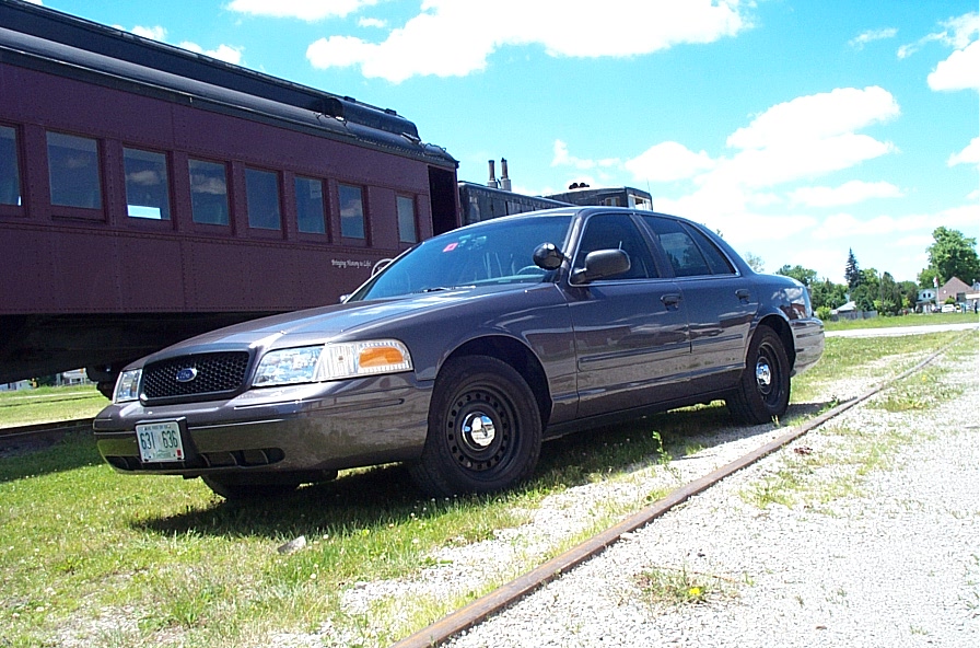 Dark gray Ford Crown Victoria parked next to a train car on a sunny day