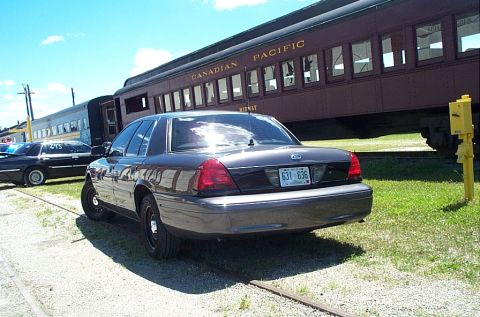 2001 Ford Crown Victoria - Rear three-quarter view of a dark gray Ford Crown Victoria sedan on black steel wheels parked on grass beside a maroon Canadian Pacific pas…