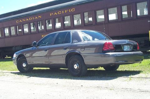 2001 Ford Crown Victoria - A gray/taupe Ford Crown Victoria sedan viewed from the rear driver's side, parked on gravel with black steel wheels and no hubcaps. Behind…
