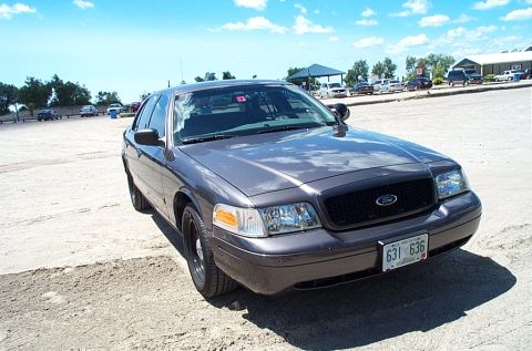 2001 Ford Crown Victoria - Grey Ford Crown Victoria Police Interceptor sedan parked on a dirt lot, shot from the front three-quarter angle with black steel wheels, bl…