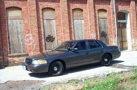 2001 Ford Crown Victoria - Grey Ford Crown Victoria Police Interceptor photographed from the driver's side alongside an abandoned red brick building with boarded-up a…
