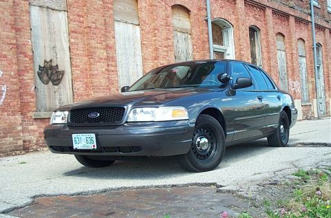 2001 Ford Crown Victoria - Low front three-quarter shot of a grey unmarked Ford Crown Victoria Police Interceptor on broken pavement beside a derelict red brick build…
