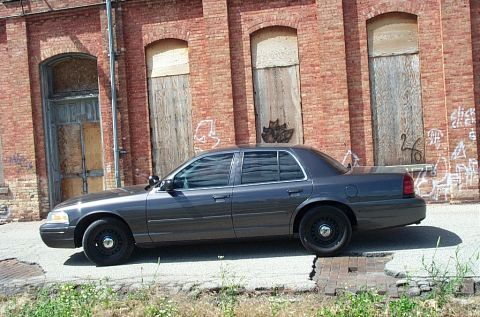 2001 Ford Crown Victoria - Side view of a dark gray 2001 Ford Crown Victoria sedan parked in front of a weathered brick building with boarded-up windows.