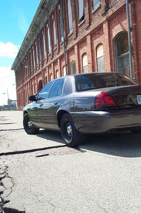 2001 Ford Crown Victoria - A dark gray Ford Crown Victoria is parked on cracked pavement in front of an old brick building.