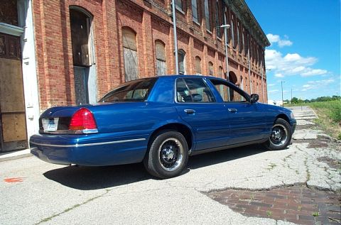 2001 Ford Crown Victoria - A blue 2001 Ford Crown Victoria sedan is parked on a cracked asphalt surface in front of a brick building.