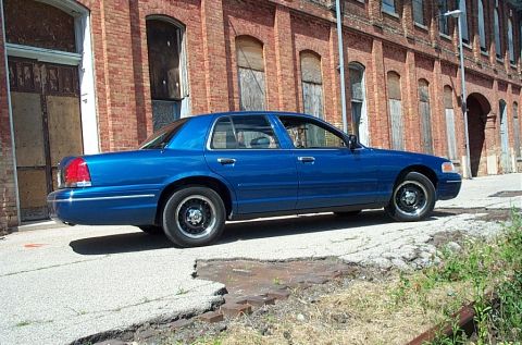 2001 Ford Crown Victoria - A blue 2001 Ford Crown Victoria is parked on a broken asphalt surface next to an old brick building.