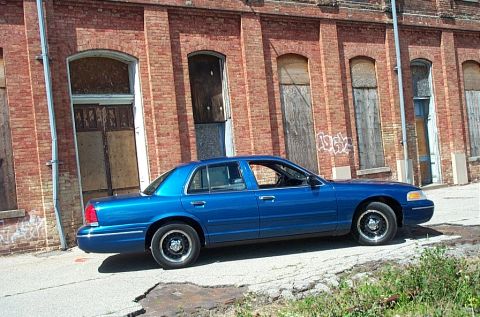 2001 Ford Crown Victoria - A blue 2001 Ford Crown Victoria sedan is parked on a cracked asphalt surface in front of a brick building with boarded-up windows.