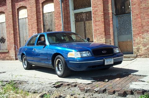 2001 Ford Crown Victoria - Blue 2001 Ford Crown Victoria parked on cracked pavement near a brick building.