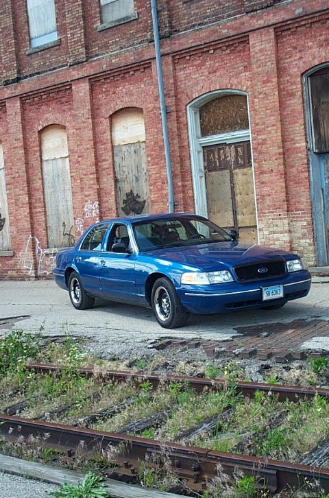 2001 Ford Crown Victoria - A blue 2001 Ford Crown Victoria is parked in front of an old brick building with boarded-up windows.